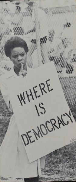 This is a 1960s black-and-white photo of an African American woman holding a protest sign that reads “Where is democracy?”